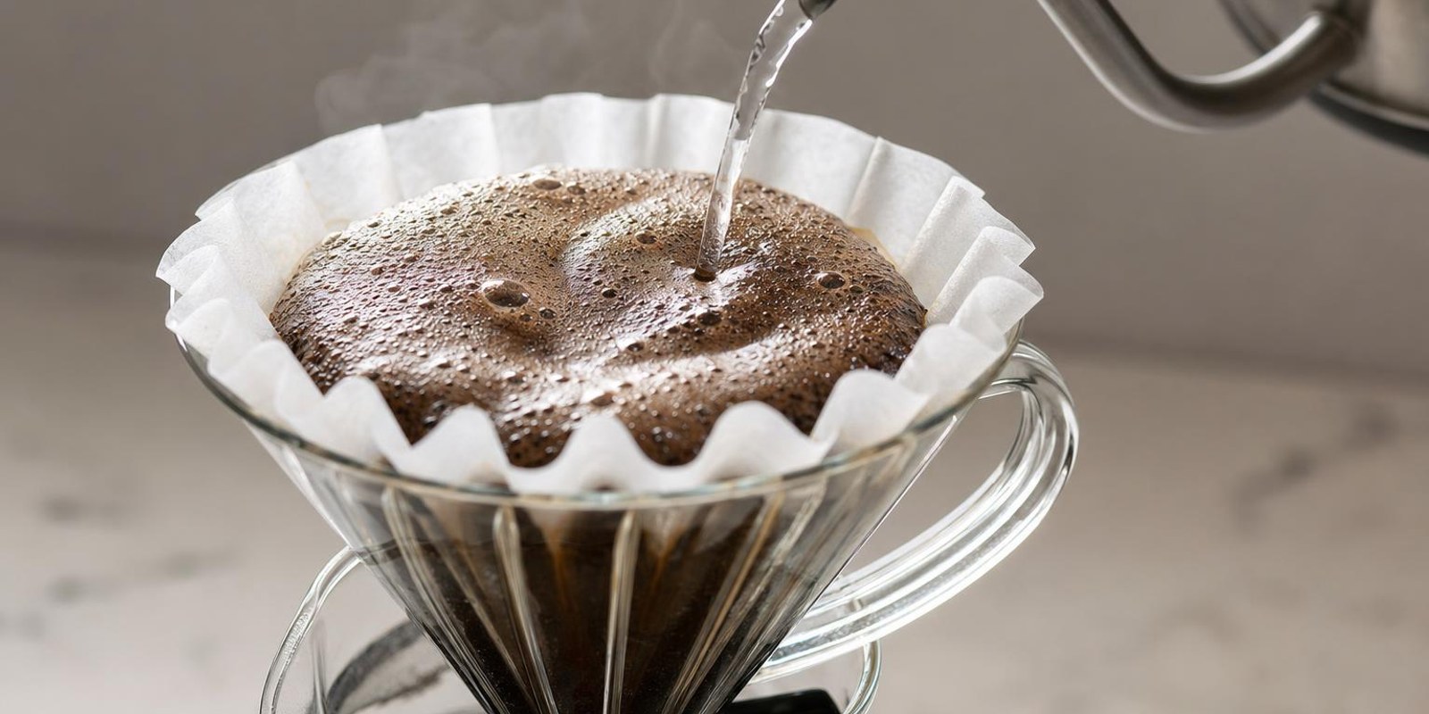 Wasser aus einer Schwanenhalskanne trifft kreisend auf aufquellendes Kaffeemehl im Handfilter.