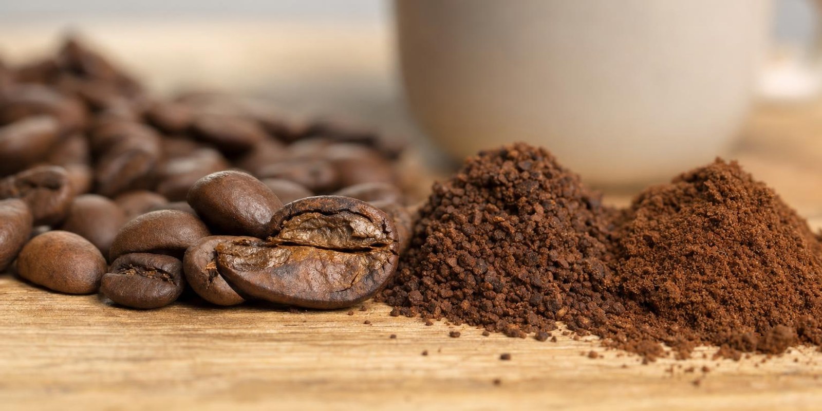 Geroestete Kaffeebohnen und unterschiedlich fein gemahlenes Kaffeepulver liegen vor einer Tasse Kaffee.