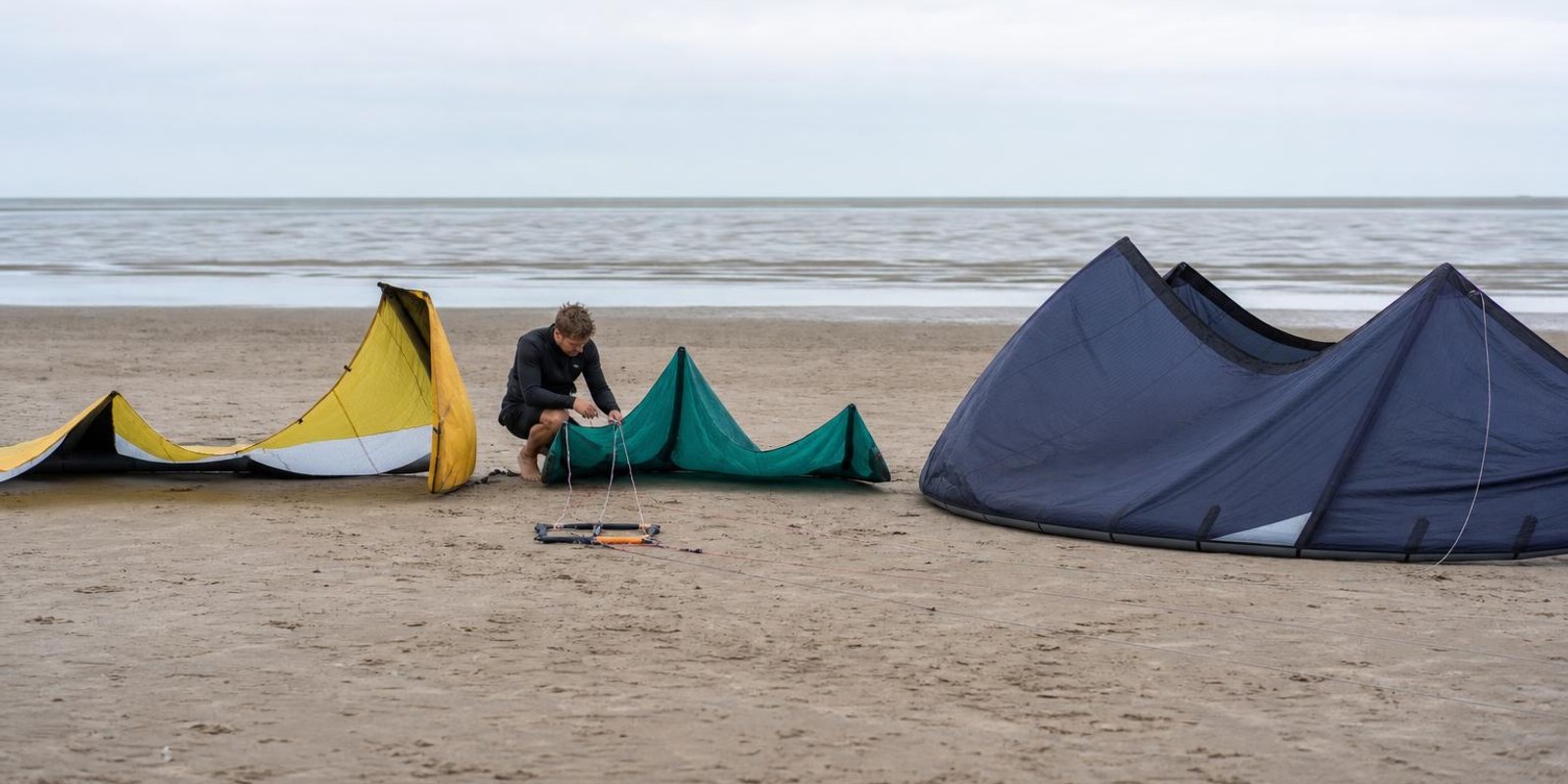 Drei unterschiedlich grosse Kites liegen nebeneinander am Strand, ein Kiter prueft die Leinen.