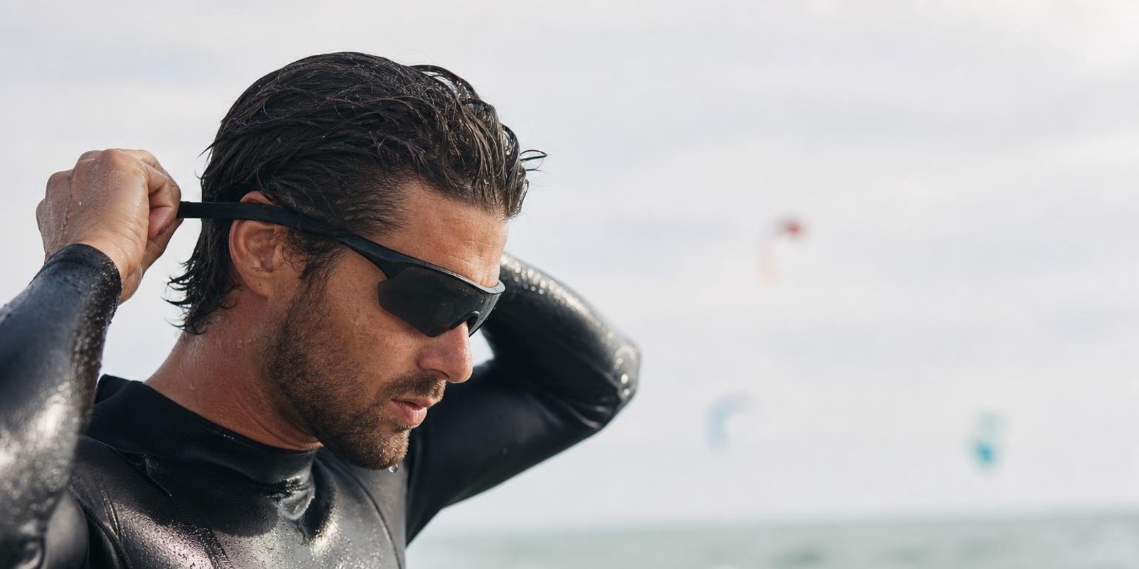 Kitesurfer befestigt eine Sportbrille mit Band, im Hintergrund Kites ueber dem Meer.