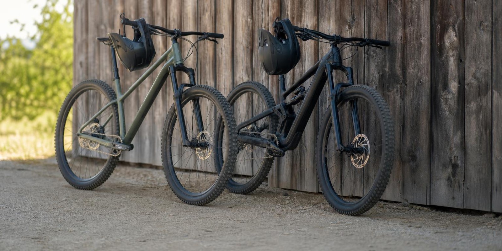 Hardtail und Fully lehnen zum Vergleich nebeneinander an einer Holzwand, Helme am Lenker.