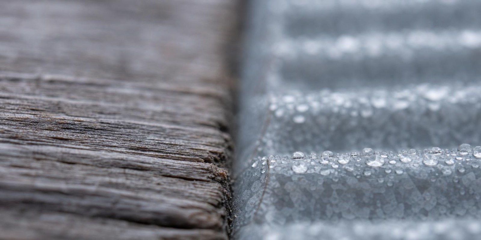 Nahaufnahme einer verwitterten Holzbohle neben einer verzinkten Metallwand mit Tautropfen.