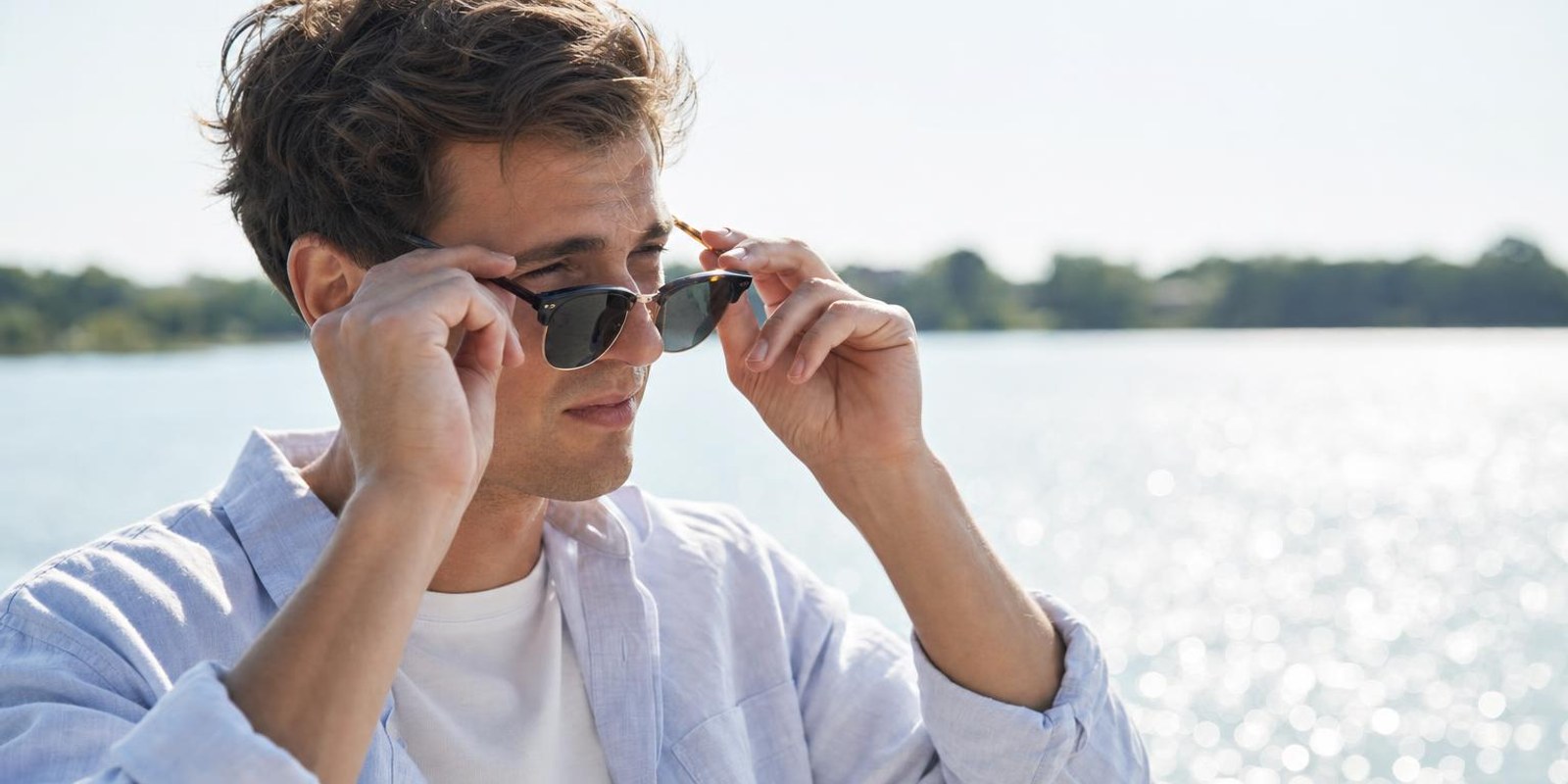 Ein Mann setzt an einem glitzernden See bei starkem Sonnenlicht eine Sonnenbrille auf.