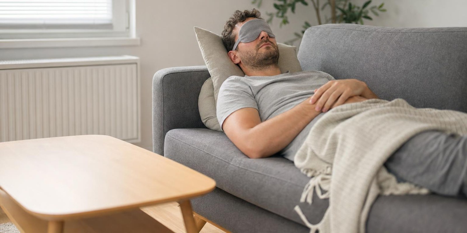 Ein Mann haelt mit Schlafmaske und leichter Decke einen kurzen Mittagsschlaf auf dem Sofa.