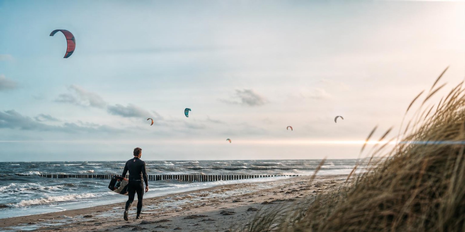 Kitesurfer traegt sein Board am Ostseestrand mit Buhnen entlang, Kites am Himmel.